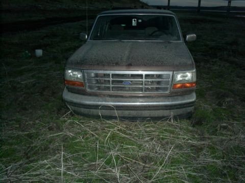1994 Ford Bronco - A 1994 Ford Bronco sits in a field, its front grille and headlights visible through a layer of dust.