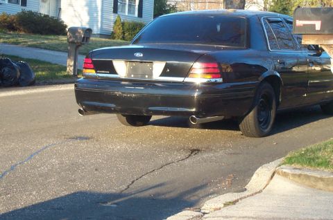 1999 Ford Crown Victoria - Black Ford Crown Victoria sedan viewed from a low three-quarter rear angle in a residential driveway, showing dual chrome exhaust tips unde…