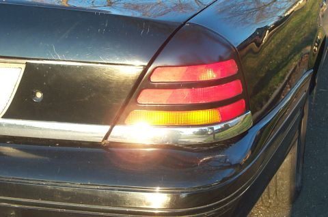 1999 Ford Crown Victoria - Sunlit close-up of the driver-side taillight on a black full-size sedan, with stacked red lenses over a lit amber turn-signal section, chro…