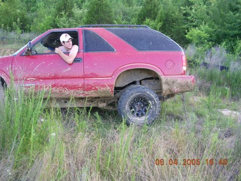 1989 Ford Bronco II - A red SUV, heavily mud-splattered, is seen from the side with a person leaning out the driver's window in a grassy, wooded environment.