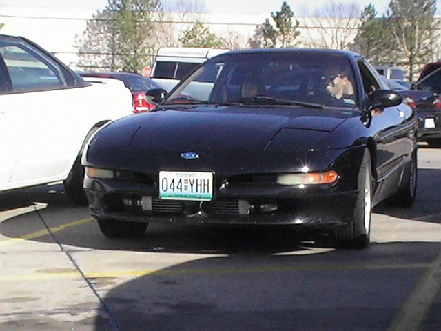 Black Ford Probe coupe with closed pop-up headlights driving through a parking lot