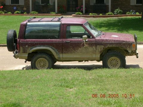 1989 Ford Bronco II - Side view of a muddy 1989 Ford Bronco II with a roof rack and spare tire mounted externally.