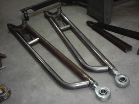 1978 Ford Bronco - A matched pair of shop-built tubular radius arms in bare welded steel lying on a shop floor, each with a forked axle-end bracket, a bracing…