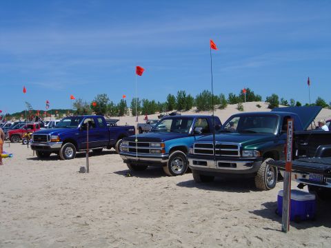 1991 Ford Bronco - Pickup trucks are parked on sand dunes under a clear blue sky with red flags flying.