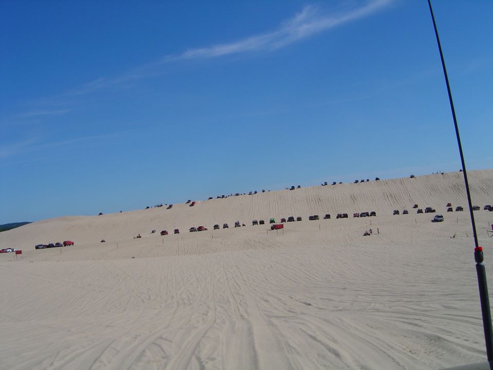 Distant line of vehicles parked along a massive sand dune ridge under blue sky
