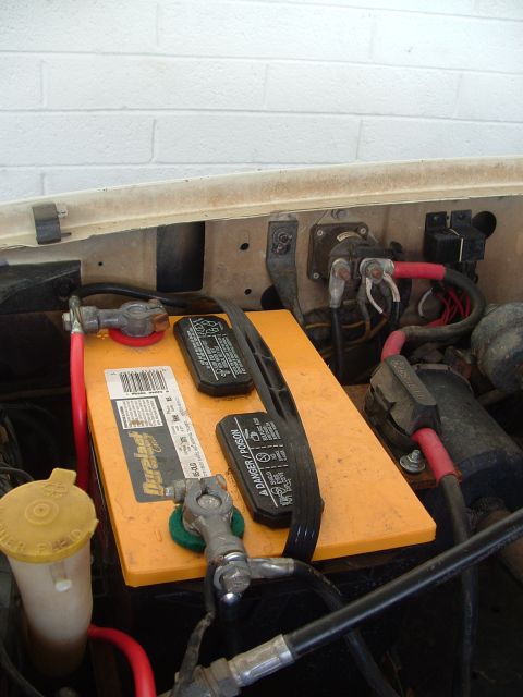 1990 Ford Bronco - Top-down close-up of a yellow Duralast battery seated in the engine bay with heavy-gauge cables on lead terminal clamps, red and green anti…