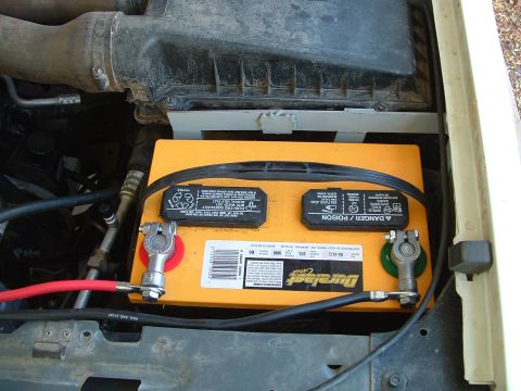 1990 Ford Bronco - Overhead shot of an orange Duralast Gold battery seated in the engine bay tray, with lead top-post terminals, red and green anti-corrosion…