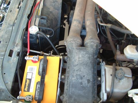 1990 Ford Bronco - Overhead view into a dusty engine bay showing a yellow-cased Duralast battery newly mounted at the driver's-side inner fender, with a red p…