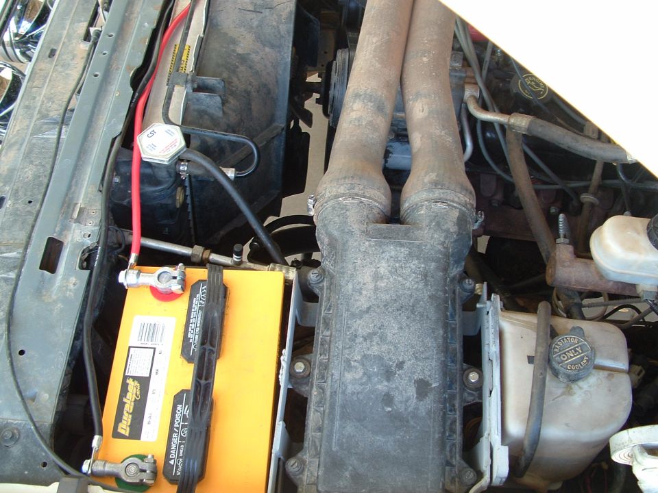Yellow Duralast battery installed in dusty engine bay beside plastic air intake tubes and coolant bottle