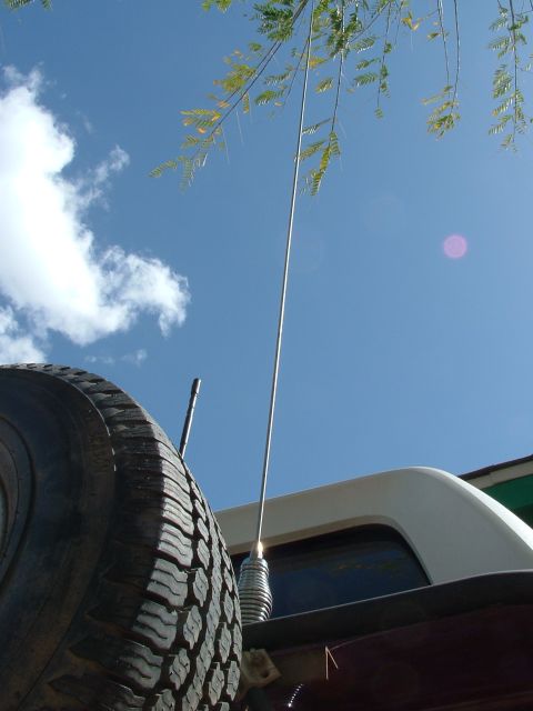1990 Ford Bronco - A spare tire and a tall antenna are visible on the rear of a vehicle under a bright blue sky.