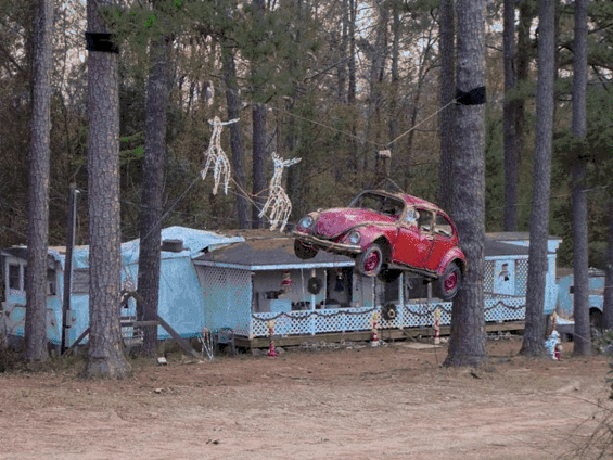 Red car being transported on a trailer, trees and a light blue structure in background