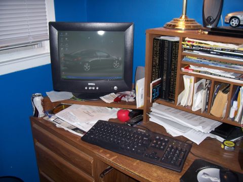 1999 Ford Crown Victoria - A cluttered desk features a computer displaying a car image, a keyboard, and various documents on a bookshelf.