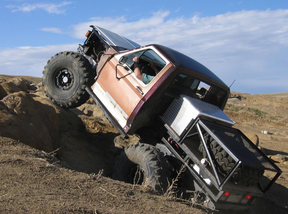 Truck with big tires going over rocks. Hood is open, showing engine. Has a custom bed rack