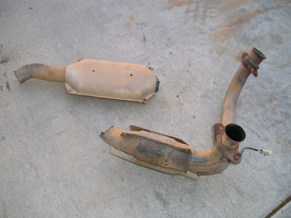 Two rusty exhaust system parts, one catalytic converter and one pipe section, lying on a concrete surface
