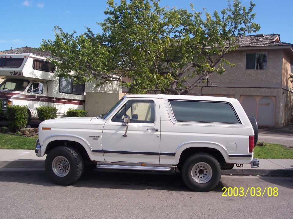 White full-size Ford Bronco, driver-side view, parked at a residential curb with an RV behind it.