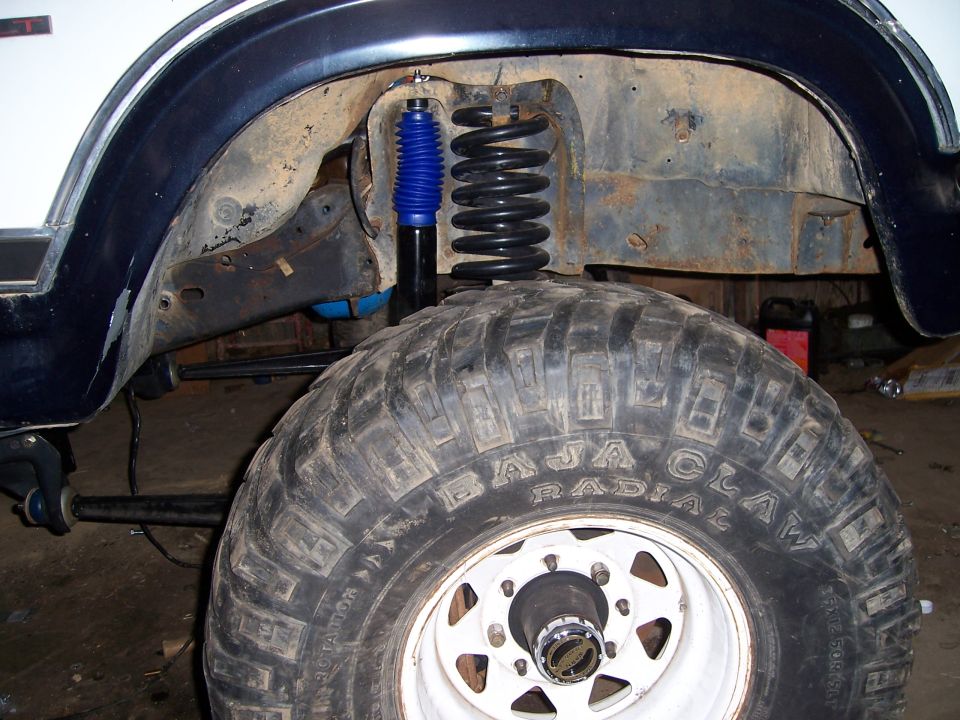 Close-up of front wheel well showing suspension, coil spring, and blue shock absorber. Baja Claw radial tire on white rim
