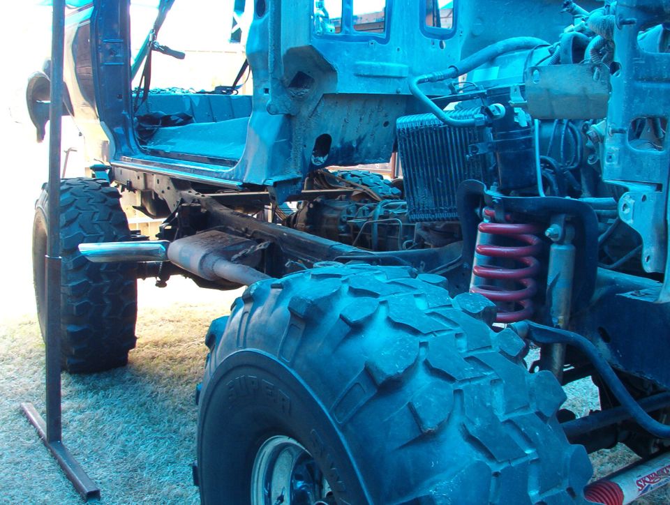 Stripped 4x4 chassis on grass with big mud tires, dual exhaust and red coil spring, blue-tinted photo