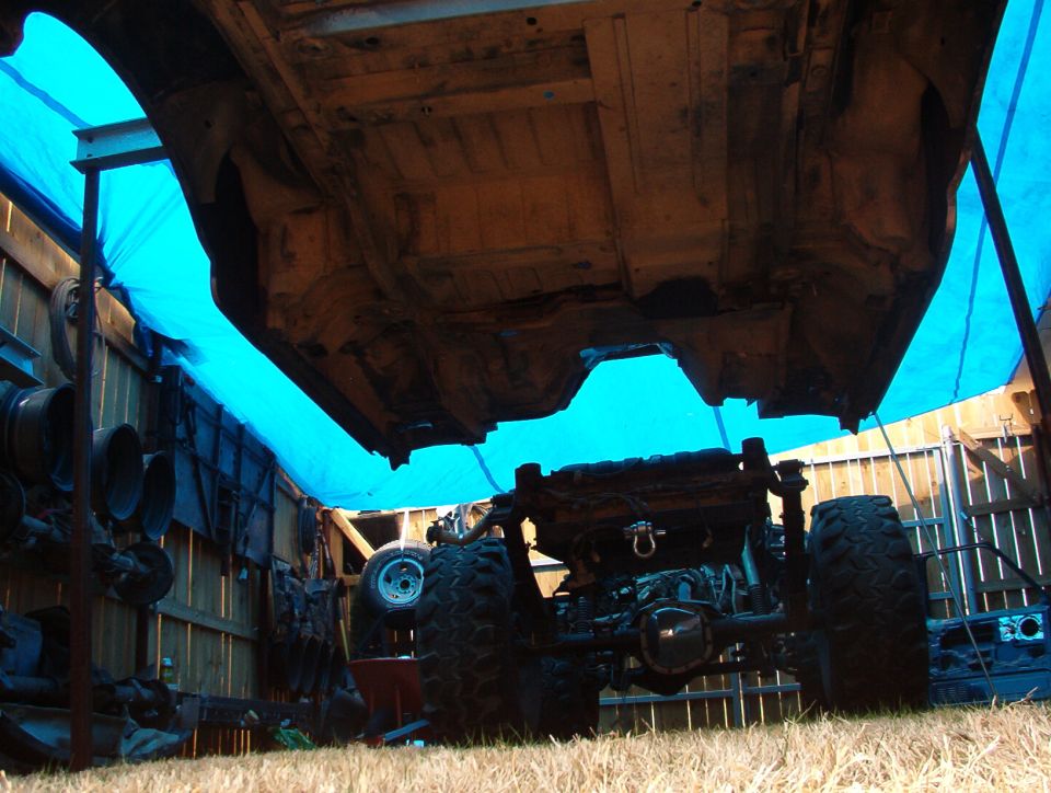 Underside of vehicle body lifted off frame with chassis and off-road tires below, under blue tarp