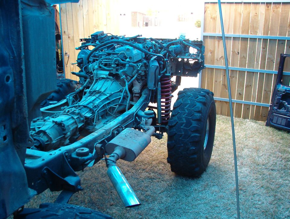 Stripped truck frame with engine, exhaust, coil suspension and off-road tire on grass by a fence