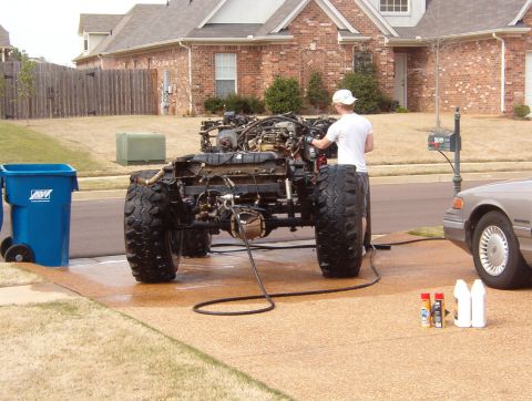 1994 Ford Bronco - A person works on a vehicle chassis in a driveway, with another car parked to the side.