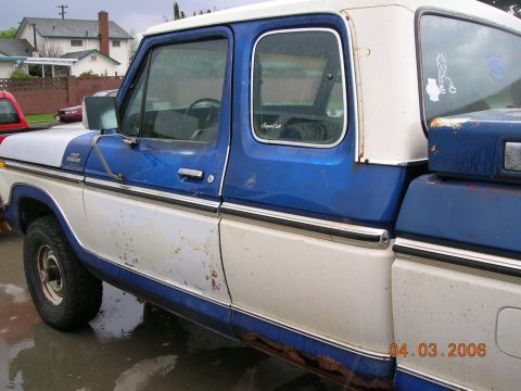 1985 Ford Bronco - This blue and white Ford F-150 pickup truck displays significant rust and wear on its body panels.