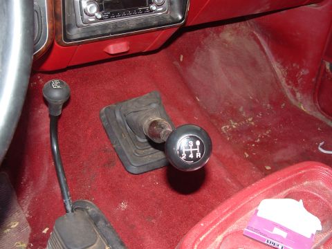 1990 Ford Bronco - Interior floor view of a vehicle with red carpet showing two black shifters: a taller manual transmission shifter with a black knob labeled…