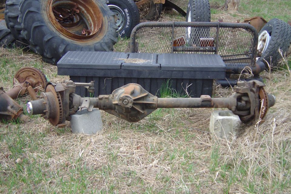Solid rear axle assembly with driveshaft and brake drums laid out on grass near old tractor tires