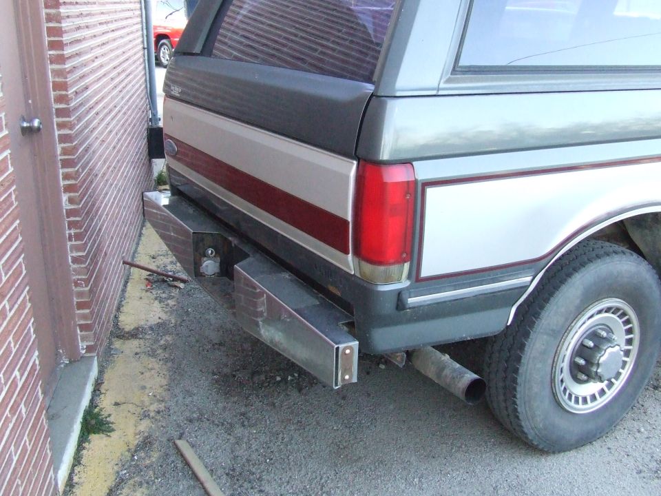 Rear view of a two-tone pickup truck with a custom bumper and camper shell. Visible taillight and wheel
