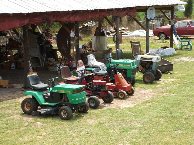 Several lawnmowers parked under a covered area with a girl sitting on one