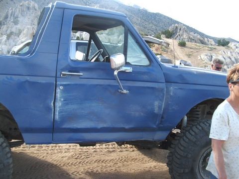 1989 Ford Bronco - A blue SUV sits parked on sandy terrain, with a mountainous landscape behind it.