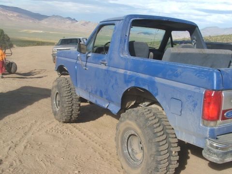 1989 Ford Bronco - A blue Ford Bronco with off-road tires is parked in a sandy area with mountains in the background.