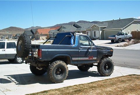 1986 Ford Bronco - This modified SUV features large off-road tires and a roll cage, with a second vehicle parked in the driveway behind it.
