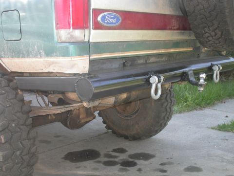1990 Ford Bronco - Rear view of a green and tan Ford SUV lifted on large off-road tires, showing a custom black tubular rear bumper with a trailer hitch ball…