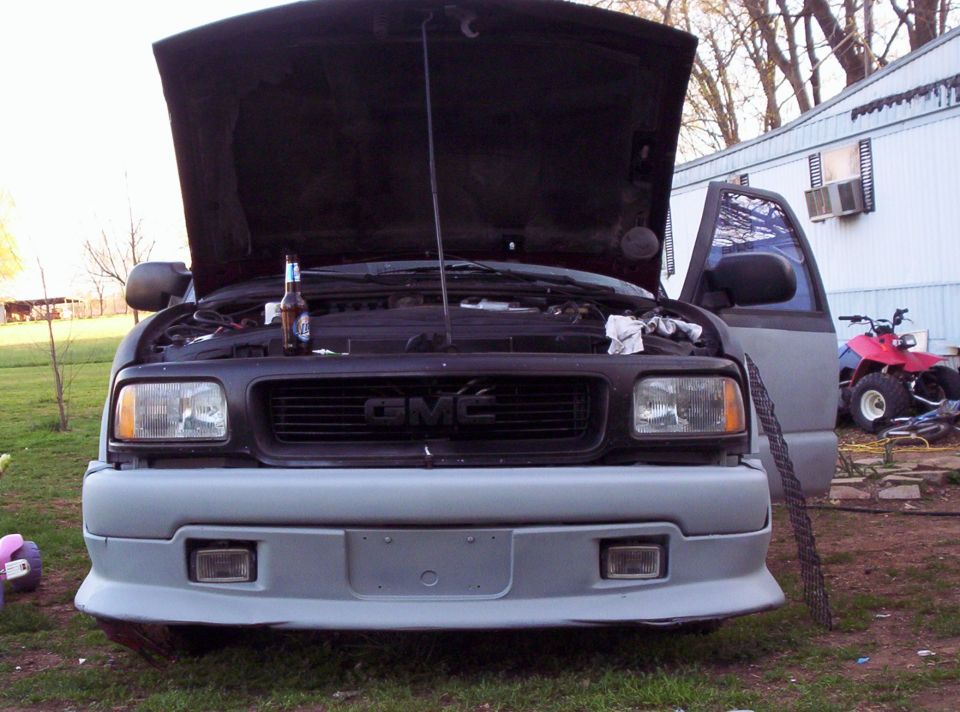 Front view of a GMC pickup truck with hood open, beer bottle on engine. ATV in background
