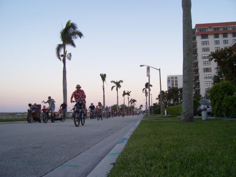 1994 Ford Probe - A group of cyclists ride along a coastal road at sunset, with palm trees and apartment buildings in the background.