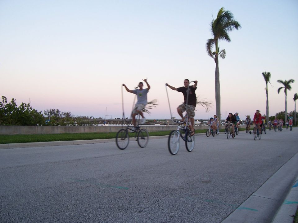 Two people ride tall bicycles with long poles, palm trees in background