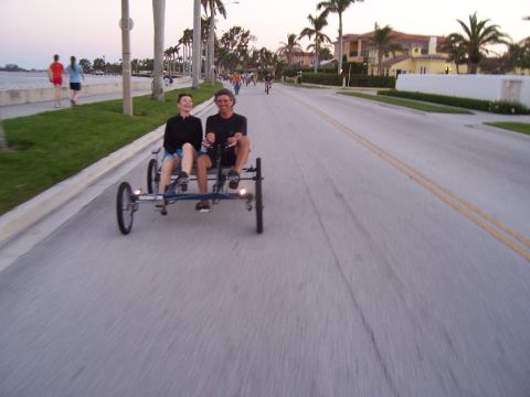 1994 Ford Probe - A quadricycle carries two riders along a coastal road at dusk, with other cyclists and pedestrians visible.