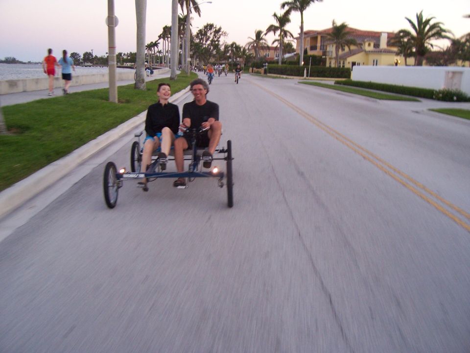 Two people ride a quadricycle on a paved road lined with palm trees and houses