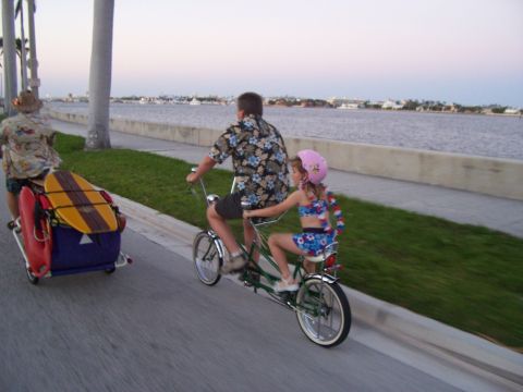 1994 Ford Probe - A father and daughter enjoy a ride on a tandem bicycle, pulling a trailer with surf gear, along a waterfront path.