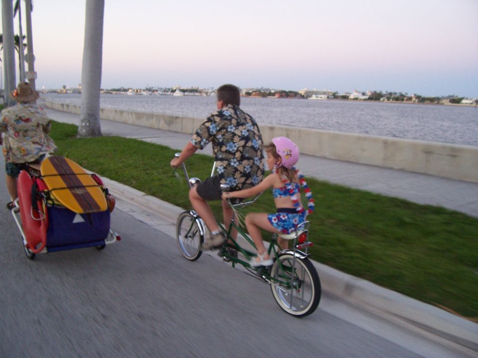 Dad and daughter ride a tandem bike past a trailer with a surfboard. Water and buildings in background
