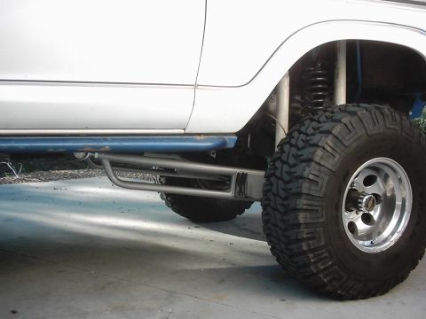1978 Ford Bronco - Low side view along the rocker of a lifted white 4x4, showing a blue tubular rock slider above a bare-steel nerf bar and a tall coil spring…