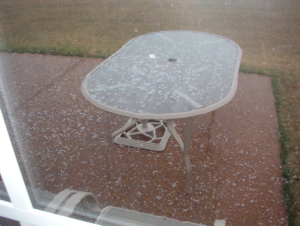 Glass-top patio table on a concrete patio dusted with small white hailstones, seen through window