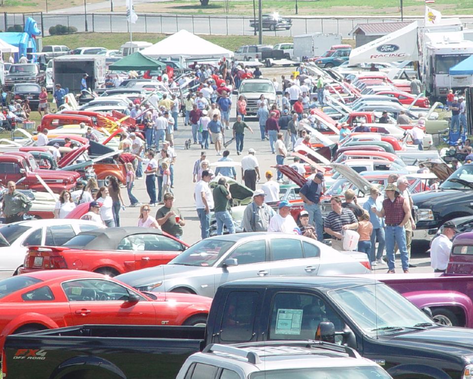 Crowded outdoor car show with rows of classics hoods-up, a Ford Racing tent, red Mustang GT and black FX4 pickup in front