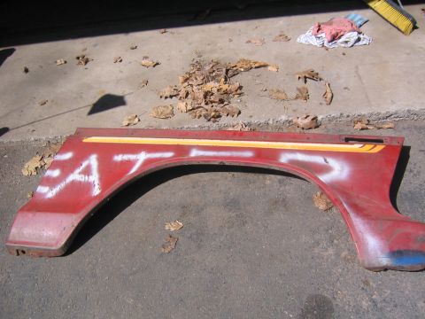 1978 Ford Bronco - A red fender with a yellow stripe and white spray-painted text lies on the ground next to fallen leaves.