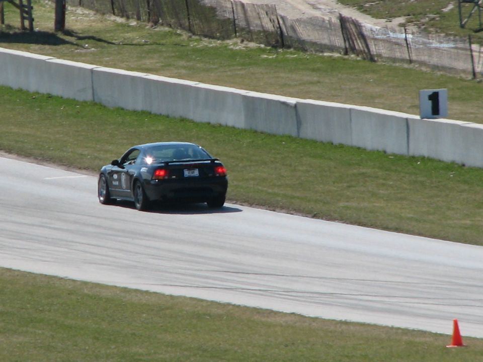 Dark coupe with spoiler cornering on a race track, rear view, near a barrier wall and number marker.