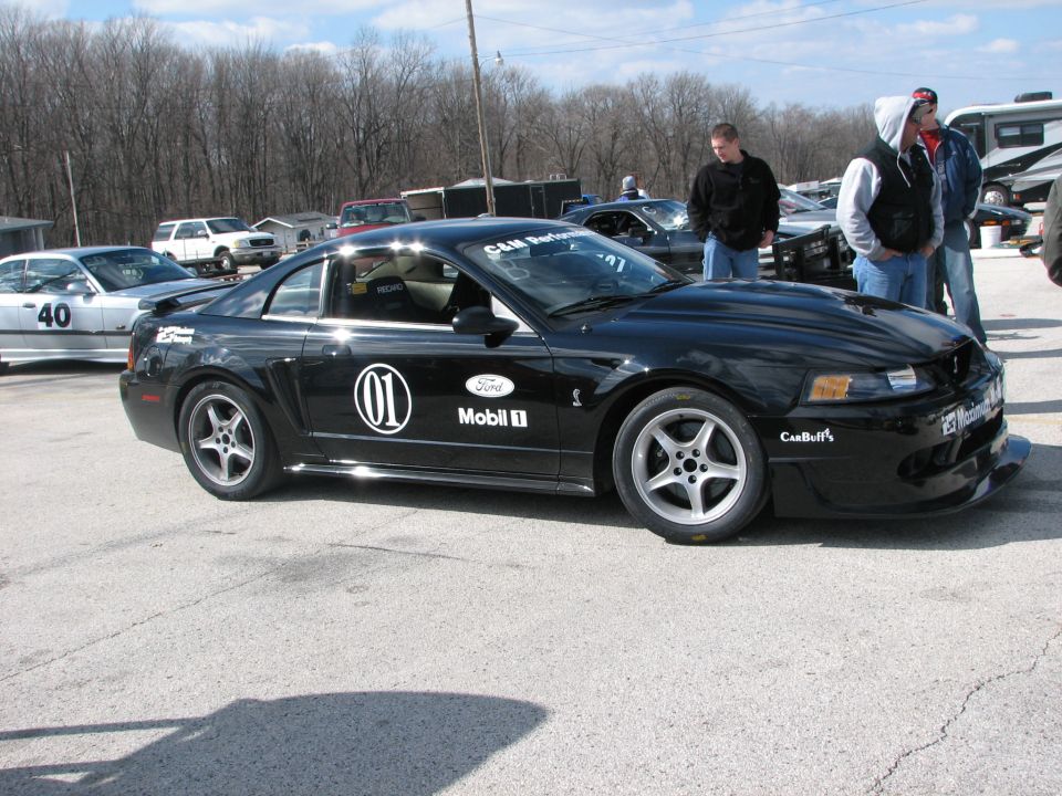 Black Ford Mustang Cobra with racing decals, parked at a track event. Another car with number 40 is visible