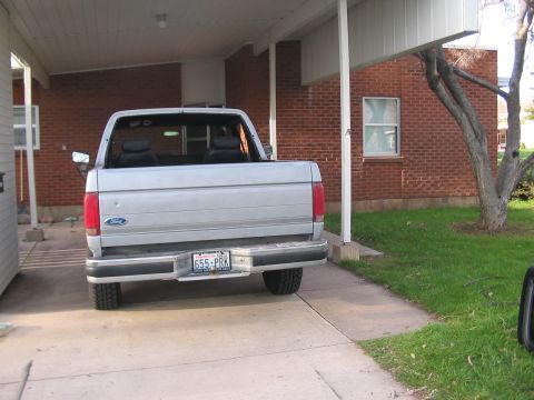 1991 Ford Bronco - The rear of a silver Ford pickup truck is visible, parked in a driveway next to a brick building.