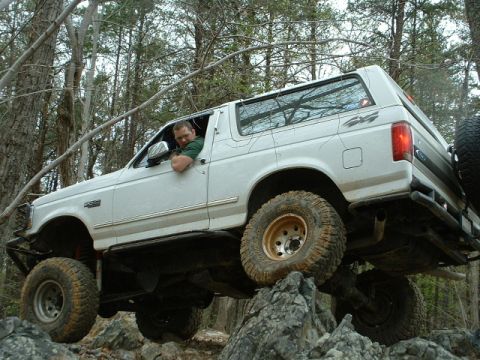 1987 Ford F-150 - Driver maneuvers a lifted white Ford Bronco over rocks on a wooded trail.