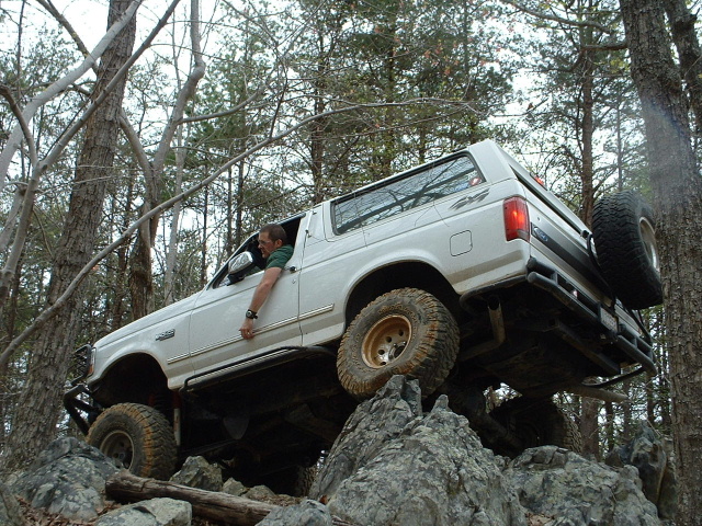 White Ford Bronco climbing over rocks on a wooded trail. Driver's arm is out the window