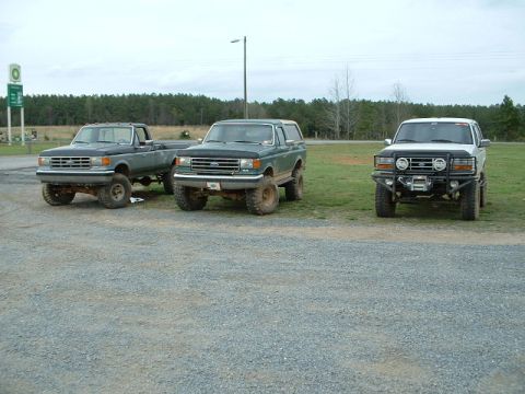 1987 Ford F-150 - Three Ford trucks and SUVs are parked on a gravel lot with a wooded background.